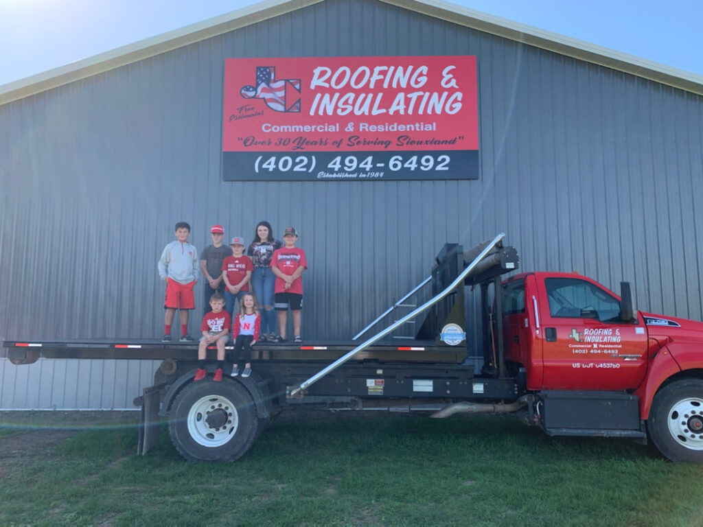 kids standing on the back of a truck in front of a warehouse with a sign for JC Roofing